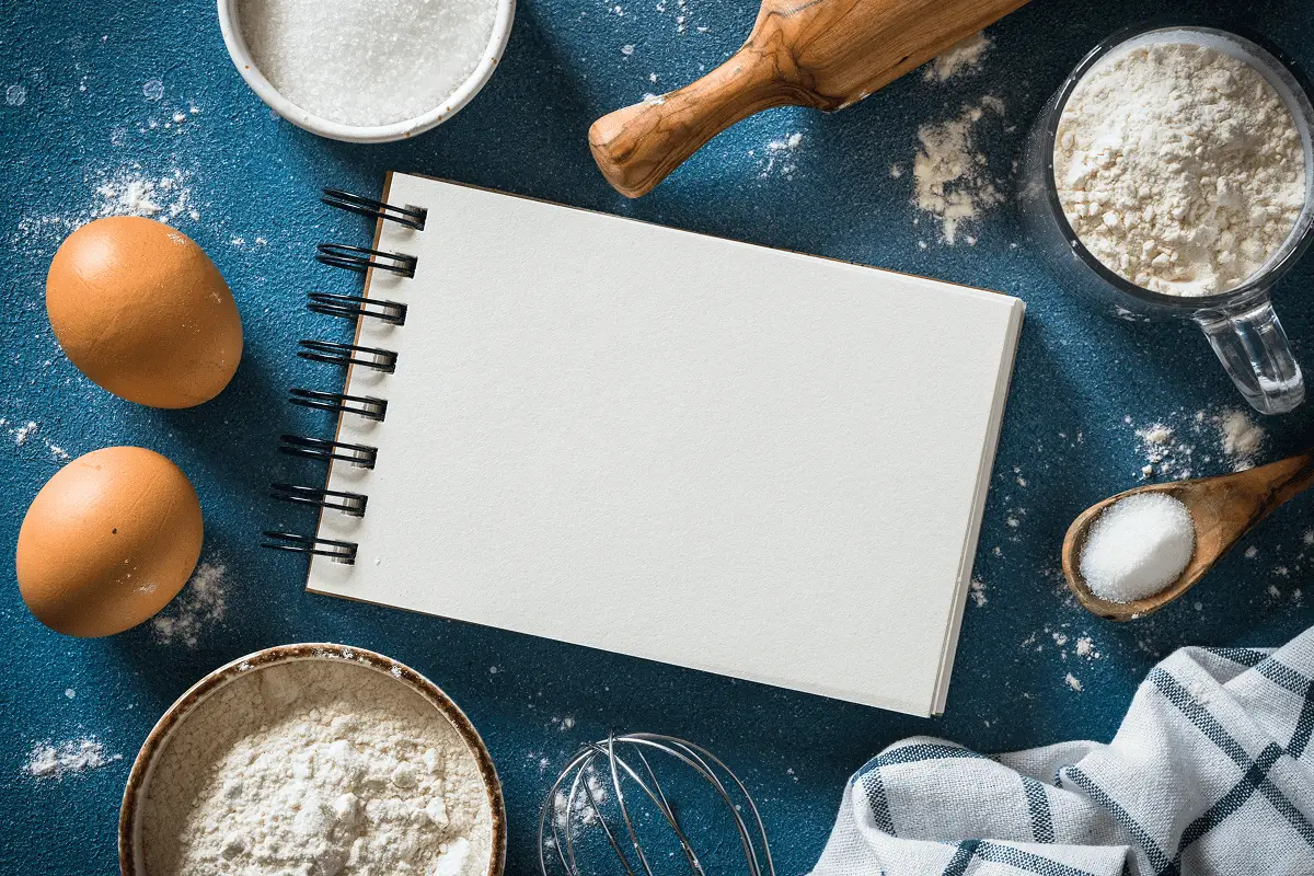Baking ingredients with blank notebook and utensils.